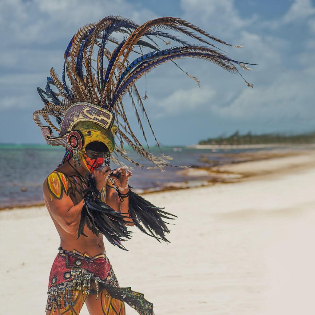 A man in tribal costume plays an instrument on a sunny Mexican beach.
